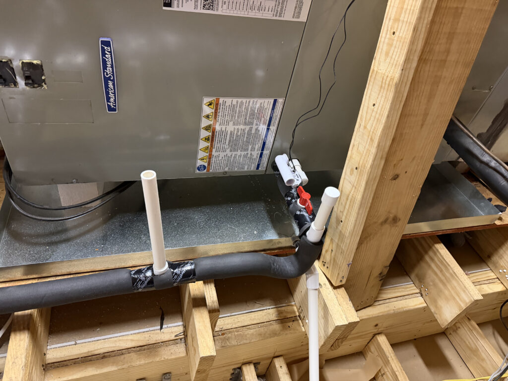 American Standard HVAC air handler unit with insulated refrigerant lines, PVC condensate drain pipes, and a red shutoff valve, mounted above exposed floor joists